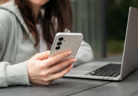 A person using a smartphone while sitting with a laptop on a wooden table outdoors, symbolizing multitasking and modern technology.の写真素材
