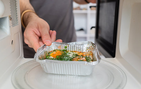 Person placing ready-to-eat meal in aluminum tray into a microwave oven for heating.の写真素材