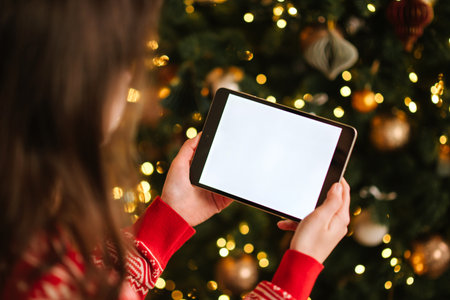 Woman holding tablet with white blank screen in front of Christmas tree decorated with lights and ornaments.の写真素材