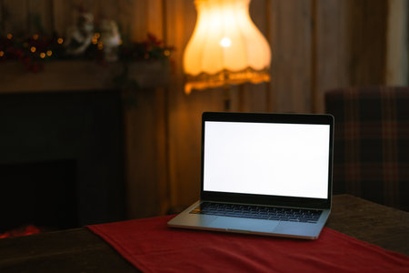 Laptop with white screen mockup on a table with a red runner in a cozy, dimly lit interior with a fireplace and vintage lamp.の写真素材