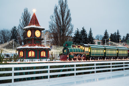 Festive Christmas village display with a toy clock tower and a green holiday train next to a decorated outdoor ice skating rinkの写真素材