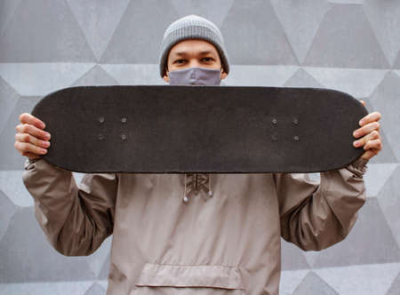 European guy in medical mask holds skateboard on his shoulder. Gray concrete wall background. Skateboarder in a gray jacket and hat. Coronavirus protection measures.の写真素材