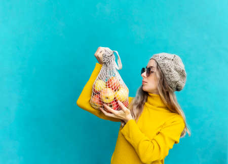 Girl holds a string bag with apples. Young woman in yellow sweater, hat and sunglasses on blue...
