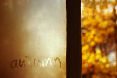 Autumn word on dirty damp window with yellow blurred leaves in the dark background and selective focus, red tonedの写真素材