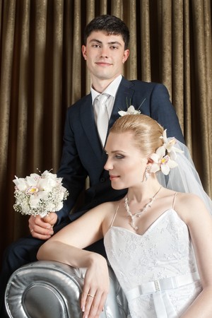 Bride with flower in hair and groom sitting behind against brown curtain foldsの写真素材