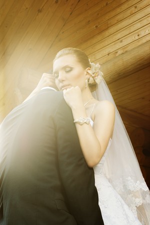 bride embracing groom with closed eyes and sunny flare against blurred wooden house logsの写真素材