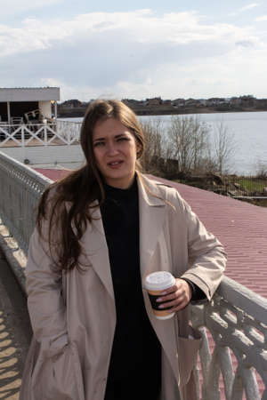 beautiful young girl with brown hair green eyes in the bright trench with coffee on the waterfront