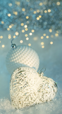 Christmas greeting postcard. A glass heart and a white ball with pearls on a snow. Beautiful blurred background of sparkling bokeh and lights of garland. Shallow depth of field.Toned image. Copy spaceの写真素材