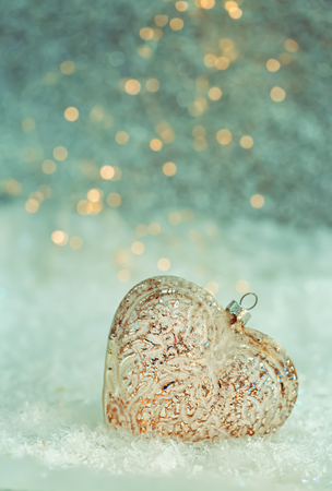 Holiday decoration. Glass heart on a snow and blurred green background of glittering bokeh with glowing lights. Happy Merry Christmas. Toned image. Copy space. Game of color. Shallow depth of field.の写真素材