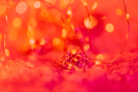 Small decorative ball with a mirror and a glittering garland on a snow. Blurred festive red background with bokeh. Happy Merry Christmas and New year. Shallow depth of field.Toned image. Copy space.の写真素材