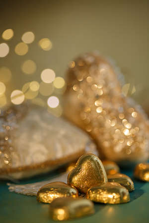 Heart shaped chocolates in golden foil. Blurred beige hearts with bokeh on a green table. Valentine's Day. Dark background. Wedding card. Shallow depth of field. Toned image. Soft focus.の写真素材