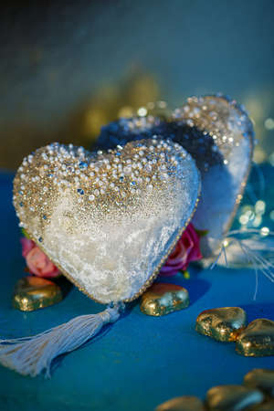 Two velvet white hearts with beads and golden foil chocolates on a blue table. Dark blurred background with bokeh. Flowers.の写真素材
