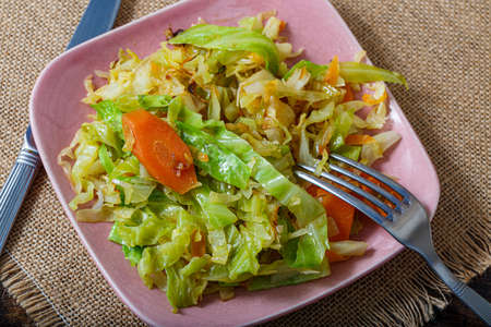 Cabbage fried or stewed with herbs, carrots, onions on a pink square plate, fork and knife on a tablecloth on the table.の写真素材