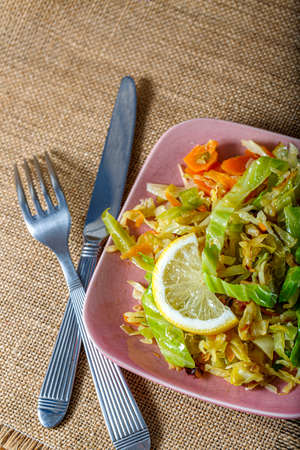 Top view of cabbage fried or stewed with herbs, carrots, onions and a slice of lemon on a pink square plate, fork and knife.の写真素材