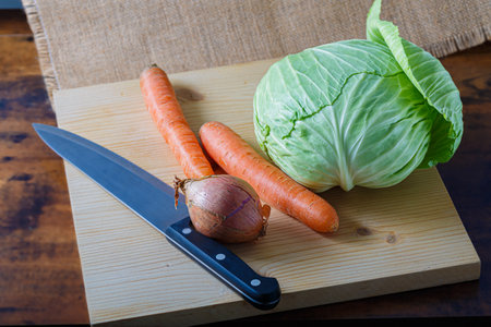 Vegetables carrots, an onion with peel and white cabbage with green leaves on a wooden board, a kitchen knife. Vegetarian food.の写真素材