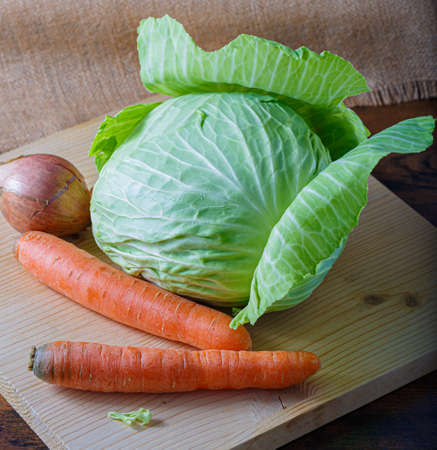 Fresh raw vegetables carrots, an onion with peel and white cabbage with green leaves on a wooden board. Vegetarian healthy food.の写真素材