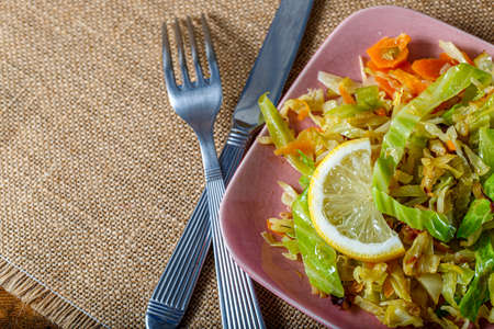 Top view of cabbage fried or stewed with herbs, carrots and a slice of lemon on a pink square plate, fork and knife.の写真素材