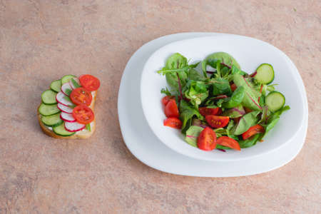 Salad of green vegetables and tomato in a white plate on the table. Spring vegetable salad with a vegetable sandwich on a stle in a white plate.の写真素材