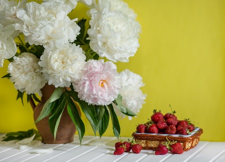 A large bouquet of peonies in a clay vase and a bowl of strawberries on a tree table. Romantic mood.の写真素材