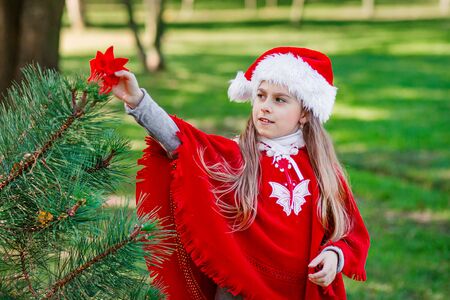 Portrait of a little girl in a Santa Claus hat near a Christmas tree in the forest.の写真素材