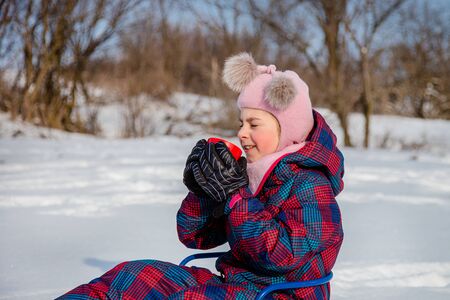 Cute girl with a red cup sits in a sleigh in the snow. The child warms himself with hot tea on a sunny winter day.の写真素材
