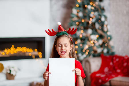 A cute girl in a Santa hat writes a letter to Santa near the Christmas tree. Happy childhood, a time for fulfilling desires. Merry Christmas.の写真素材