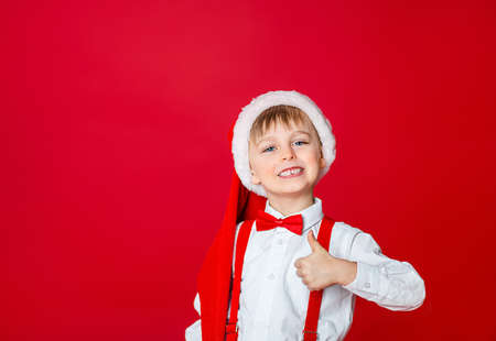 Merry Christmas. Cute cheerful little boy in Santa Claus hat on red background. A happy childhood with dreams and gifts. Close-up of baby's open mouth, milk tooth fell out.の写真素材