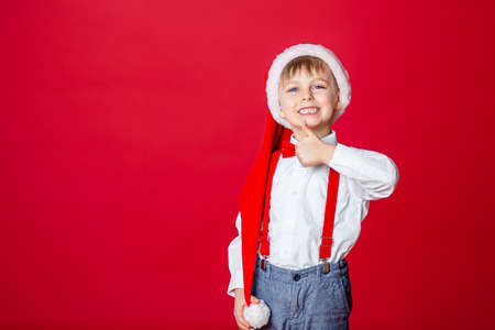Merry Christmas. Cute cheerful little boy in Santa Claus hat on red background. A happy childhood with dreams and gifts. Close-up of baby's open mouth, milk tooth fell out.の写真素材