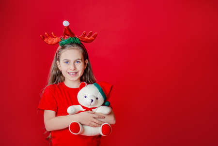 Portrait of a funny cheerful girl with a bandage of horns on her head hugging a teddy bear in Christmas pajamas isolated on a bright red background. The child points a hand, a place for text.の写真素材