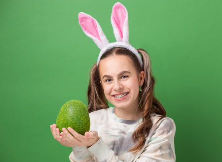 Happy Easter. Cheerful girl with Easter bunny ears and big Easter egg made of green grass on green background. Copy space. Smiling girl in braces.の写真素材