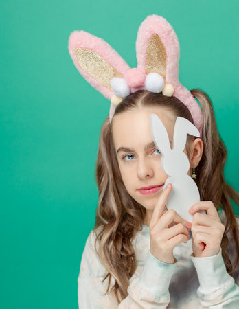 Happy Easter. Girl with happy smile, wearing shiny pink bunny ears and holding wooden bunny figurine, blue background, radiating Easter fun. Cute smile with braces on her teeth.の写真素材