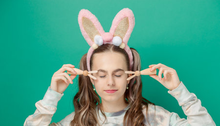 Happy Easter. Girl with joyful smile, wearing glittery pink bunny ears and holding wooden bunny figures, showcases her braces against vibrant teal background, radiating Easter fun.の写真素材