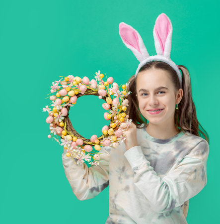 Happy Easter. Girl with joyful smile, in shiny pink rabbit ears and holding an Easter wreath of flowers, against blue background. He smiles sincerely with braces on his teeth.の写真素材