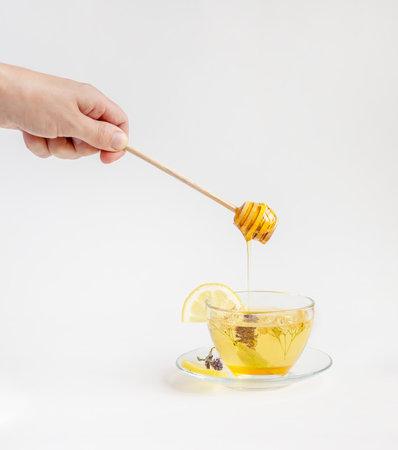 Hand dripping honey on a cup of herbal tea surrounded by flowers and bottles. Concept of healthy lifestyle and natural remediesの写真素材