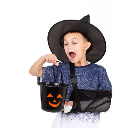 Halloween celebration concept. Portrait emotional cheerful boy in black witch hat with a hand in a cast, with bucket pumpkin Jack for sweet candies. Isolated white backgroundの写真素材