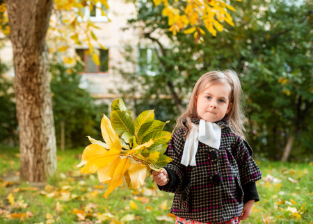 Little girl holding yellow autumn leaves, smiling in outdoor park. Seasonal joy and child happiness conceptの写真素材