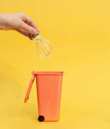 Close-up of hand placing clear light bulb into small orange trash bin against vibrant yellow background, symbolizing waste, recycling, or ideas.の写真素材