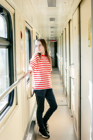 Smiling teen girl in red and white striped shirt stands inside sunlit train corridor, expressing...
