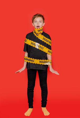 Boy stands with yellow warning tape wrapped around his body and mouth, creating a dramatic visual on bold red background.の写真素材