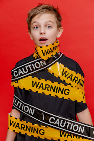 Boy stands with yellow warning tape wrapped around his body and mouth, creating a dramatic visual on bold red background.の写真素材