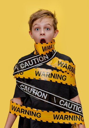 Boy stands with yellow warning tape wrapped around his body and mouth, creating a dramatic visual on bold background.の写真素材