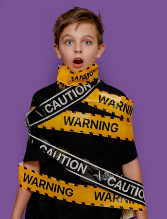 Boy stands with yellow warning tape wrapped around his body and mouth, creating a dramatic visual on bold violet background.の写真素材