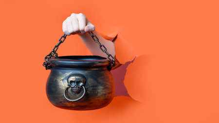 A hand holding a black Halloween cauldron with skull and chain through torn orange background.の写真素材