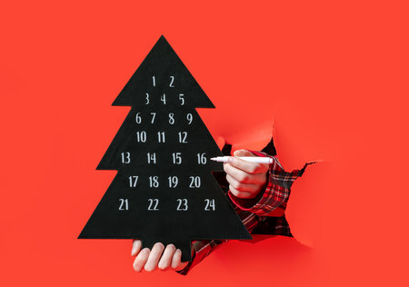 Merry Christmas. person in a plaid shirt holds a black Christmas tree-shaped advent calendar through torn red paper, marking dates with a white pen.の写真素材