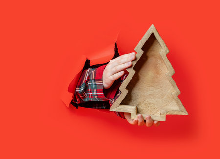 Merry Christmas. A hand in plaid shirt holds a wooden tray shaped like a Christmas tree through a torn red paper background.の写真素材