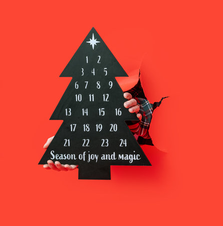 Merry Christmas. person in a plaid shirt holds a black Christmas tree-shaped advent calendar through torn red paper, marking dates with a white pen.の写真素材