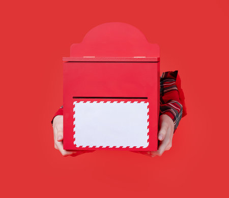 Merry Christmas, hand holds a red holiday mailbox with an envelope inserted, festive plaid shirt visible through torn paper background.の写真素材