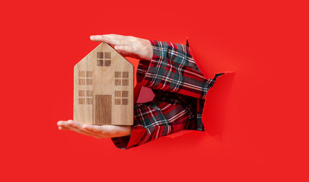 Merry Christmas. hand holding wooden toy house and pointing at it through torn red paper, plaid shirt visible.の写真素材