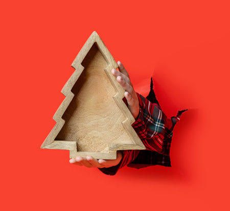 Merry Christmas. A hand in plaid shirt holds a wooden tray shaped like a Christmas tree through a torn red paper background.の写真素材