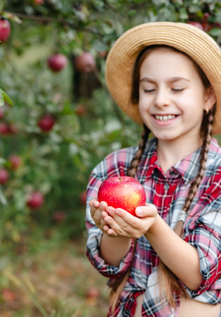 Cheerful girl stands on background an apple orchard with red apples, tastes, enjoys snacks,...
